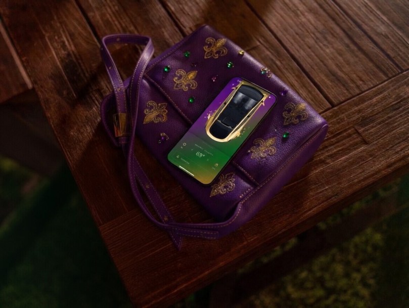 Purple Mardi Gras handbag with gold fleur-de-lis details on a wooden table, featuring a Tesla phone app showing a Tesla Model Y and 68° weather in Louisiana – creative symbol of stylish Tesla ownership in the state.