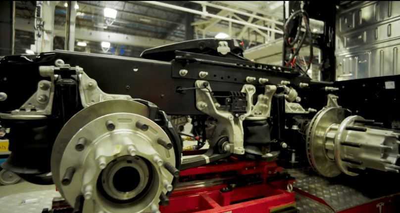 Up-close on the massive silver axle, air suspension, and heavy-duty black chassis frame of a Tesla Semi mounted on a red production rig — the raw mechanical backbone taking shape inside Tesla’s Sparks factory. Captured directly from Ashlee Vance’s Core Memory factory tour video, this provocative shot reveals the serious engineering Elon Musk is putting behind the all-electric Class 8 truck that’s poised to slash diesel pollution forever.