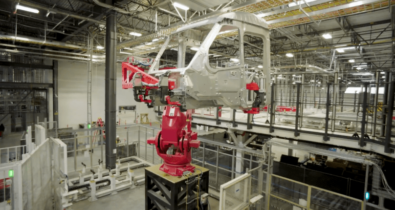 Inside Tesla’s new dedicated Semi factory in Sparks, Nevada, a large red robotic arm holds the raw, unpainted silver cab shell of a Tesla Semi high above the factory floor during the early body-in-white assembly stage. Captured directly from Ashlee Vance’s Core Memory factory tour video, this striking shot shows the bare structural framework of the all-electric Class 8 truck before painting and final assembly.