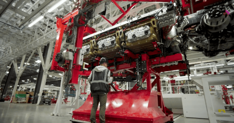 Inside Tesla’s new dedicated Semi factory in Sparks, Nevada, a production worker in a hard hat and safety vest stands beneath a massive red overhead gantry system as three large gold-colored battery packs are precisely lowered and installed into the chassis of a Tesla Semi during the critical “battery marriage” stage. Captured directly from Ashlee Vance’s Core Memory factory tour video, this shot shows the exact moment the long-range electric truck receives its power source — the same structural battery packs used in the Cybertruck.