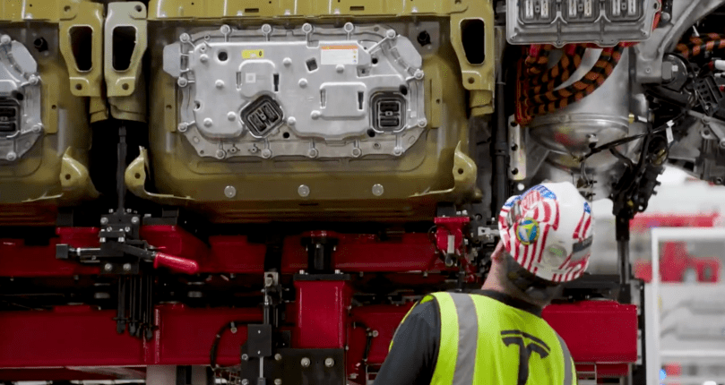 Inside Tesla’s new dedicated Semi factory in Sparks, Nevada, a proud technician in a stars-and-stripes hard hat and bright yellow Tesla safety vest watches intently as a massive gold structural battery pack — the heart of the long-range all-electric Semi — is installed into the chassis. Captured directly from Ashlee Vance’s Core Memory factory tour video.