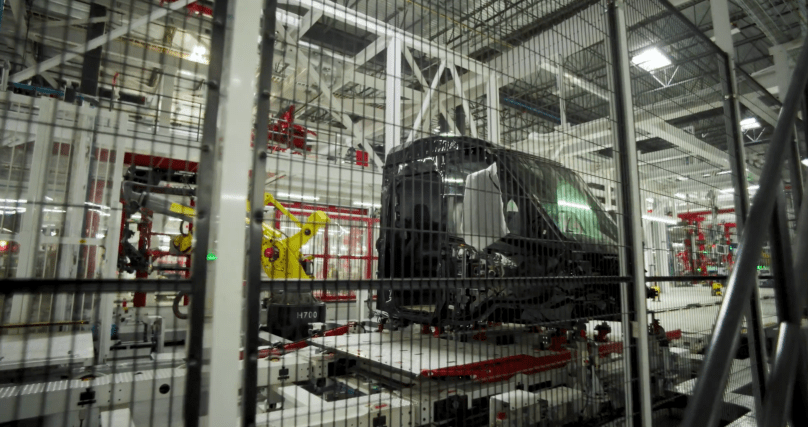 Viewed through heavy safety fencing inside Tesla’s new dedicated Semi factory in Sparks, Nevada, a glossy black Tesla Semi cab advances along the automated assembly line on a large white platform. Captured directly from Ashlee Vance’s Core Memory factory tour video, this production-stage shot shows the freshly painted truck cab — complete with protective white covering on the large windshield — as robotic arms (including the yellow H700) prepare it for the next phase of building the all-electric Class 8 semi.