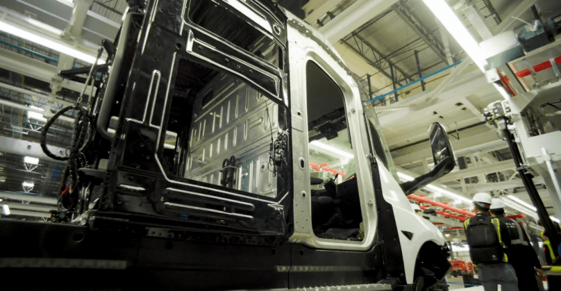 A raw, partially assembled Tesla Semi chassis stands exposed on the factory line in Sparks, Nevada — doors open, interior wiring and structural panels visible — as workers oversee the build of Elon Musk’s game-changing all-electric Class 8 truck. Captured directly from Ashlee Vance’s Core Memory factory tour video, this provocative shot reveals how close Tesla is to flooding the roads with zero-emission semis.