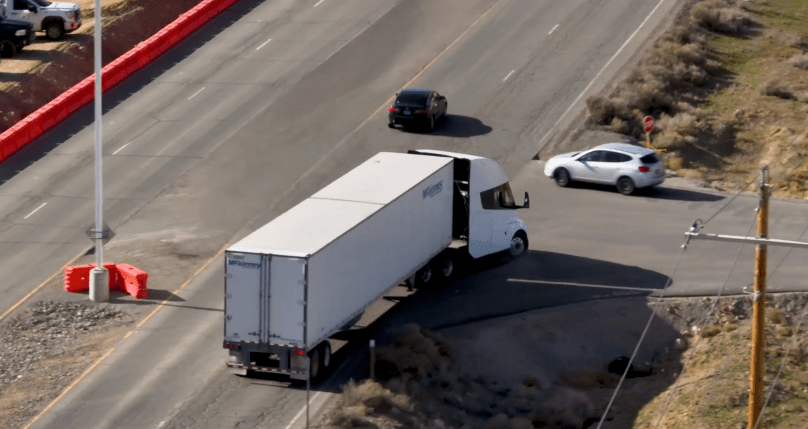 Aerial view of a production Tesla Semi electric truck in action on a Nevada highway — captured directly from Ashlee Vance’s Core Memory video. The white Tesla Semi hauls a McKinney-branded trailer alongside regular traffic, with orange construction barriers and desert scrub in the background. This real-world driving shot highlights the all-electric Class 8 semi now rolling out of Tesla’s new dedicated factory in Sparks, Nevada.