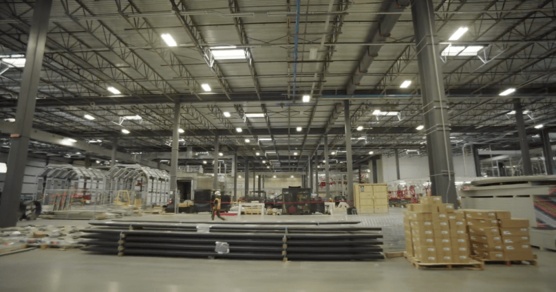 Inside Tesla’s massive new dedicated Semi factory in Sparks, Nevada, rows of unpainted silver Tesla Semi cab shells line the expansive production floor ready for assembly, surrounded by towering stacks of structural materials and parts. Captured directly from Ashlee Vance’s Core Memory factory tour video, this sweeping wide-angle shot reveals the enormous scale of Tesla’s all-electric truck production ramp-up.