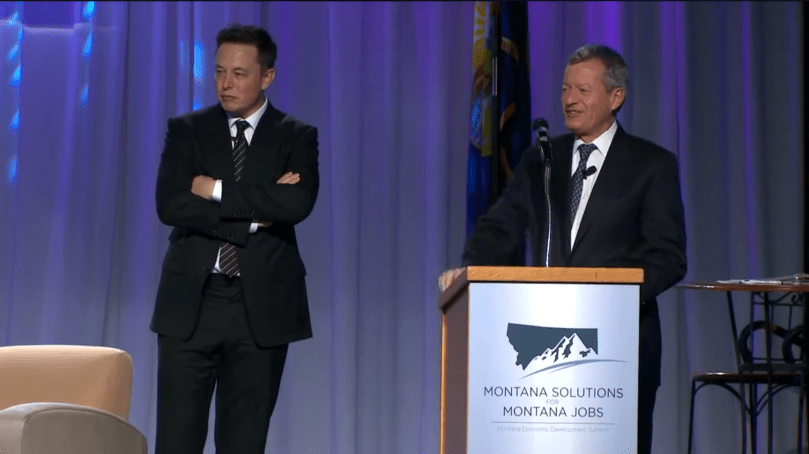 Elon Musk stands with arms crossed beside Senator Max Baucus at the 2013 Montana Economic Development Summit in Butte, Montana.